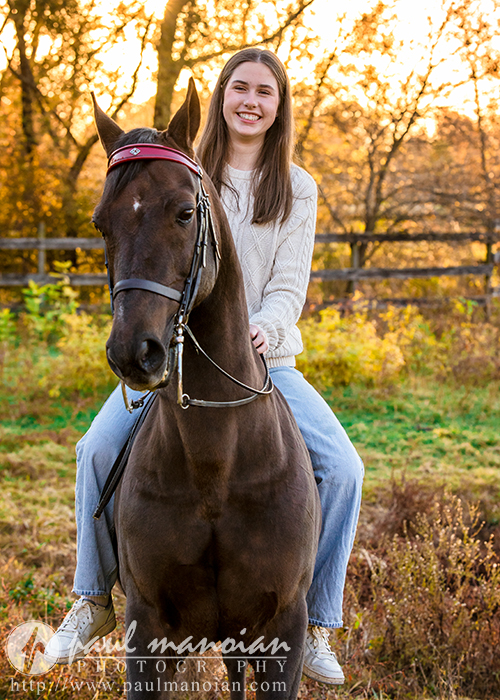 In a captivating display of photography, a high school senior in a white sweater and jeans sits on a dark brown horse. The sunlit autumn landscape, framed by trees and a wooden fence, creates the perfect backdrop for unforgettable senior pictures.