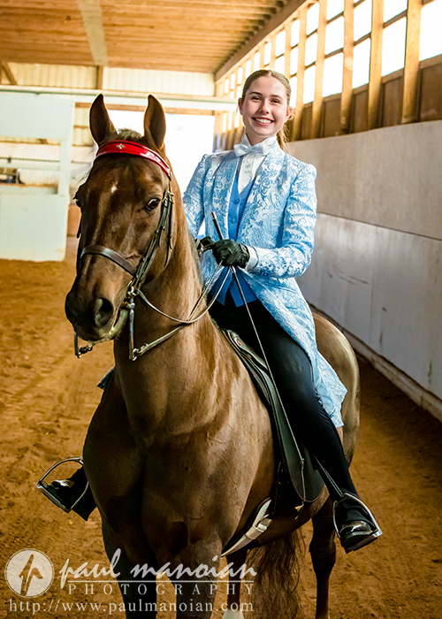 A high school senior in a blue ornate jacket and gloves is beaming while riding a brown horse inside an indoor arena. The horse, adorned with a red ribbon, looks majestic as sunlight filters through the windows above—the perfect setting for unforgettable senior portraits.