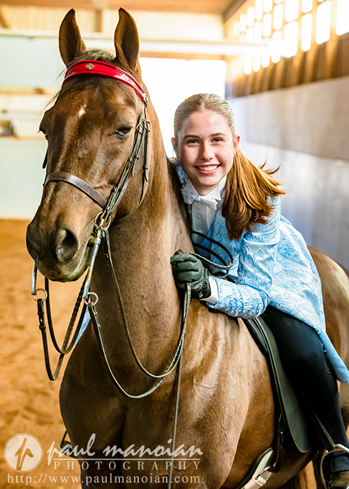 A high school senior in a blue show jacket and gloves smiles while sitting on a brown horse in an indoor arena, capturing the essence of senior photography. The horse, adorned with a bridle featuring a red browband, stands poised as sunlight filters through the windows, illuminating this perfect moment.