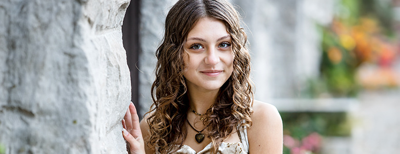 A high school senior girl with curly hair and a necklace stands outdoors, smiling gently, capturing the essence of Livonia senior pictures. They are leaning against a stone wall, with greenery and colorful flowers blurred in the background.