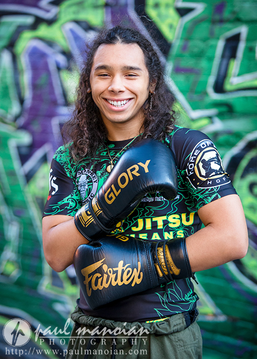A high school senior with long hair smiles confidently, wearing black and gold boxing gloves and a vibrant sports shirt. Beside them lies a skateboard, adding to the dynamic scene set against a colorful graffiti-covered wall—an ideal backdrop for unique senior pictures.