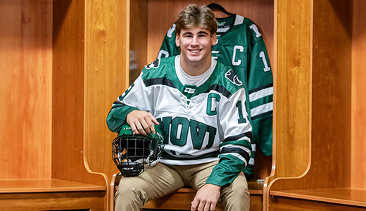 A high school senior hockey player grins in the locker room, wearing a green and white jersey for his Novi senior pictures. With a helmet in hand and a matching jersey hanging behind him, he sits beneath the warm glow of wooden lockers.