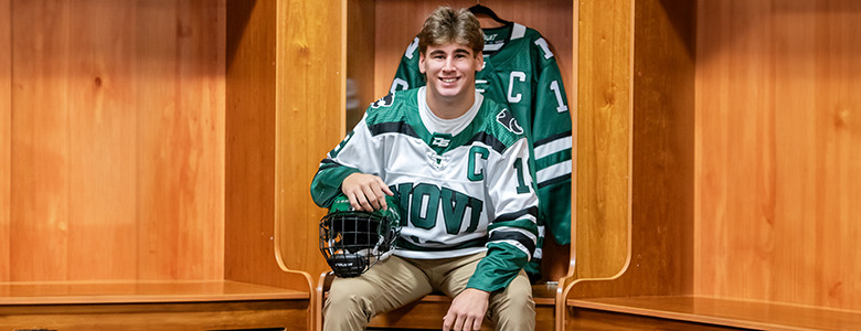 A high school senior hockey player grins in the locker room, wearing a green and white jersey for his Novi senior pictures. With a helmet in hand and a matching jersey hanging behind him, he sits beneath the warm glow of wooden lockers.