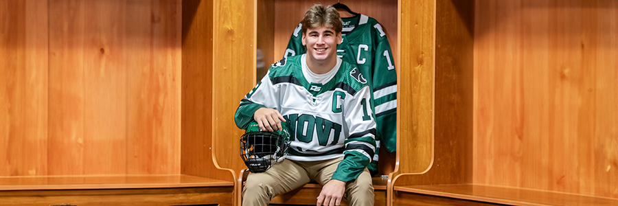A high school senior hockey player grins in the locker room, wearing a green and white jersey for his Novi senior pictures. With a helmet in hand and a matching jersey hanging behind him, he sits beneath the warm glow of wooden lockers.