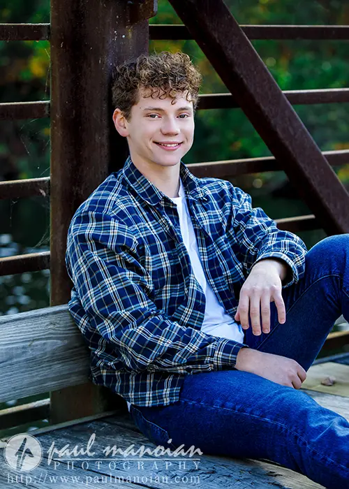 A high school senior with curly hair sits on a wooden bridge railing in Ann Arbor, smiling. They're wearing a plaid shirt over a white t-shirt and blue jeans. The background is a blurred view of trees and the river, capturing the perfect moment for senior pictures.