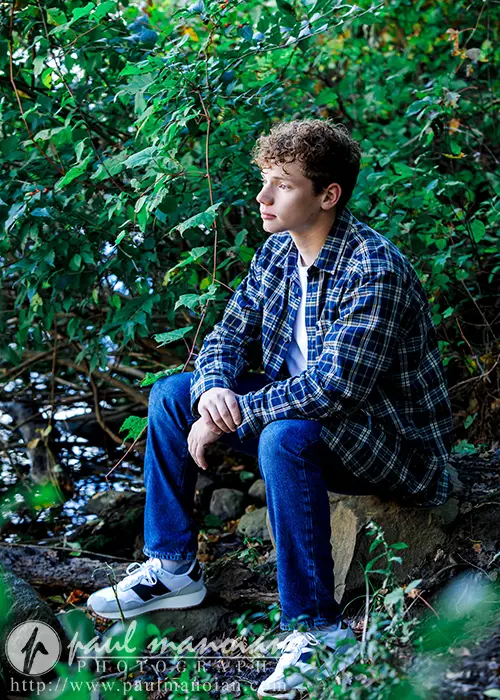 A high school senior with curly hair sits on a rock near a small pond, surrounded by lush Ann Arbor greenery. Wearing a plaid shirt, jeans, and sneakers, he gazes thoughtfully into the distance—capturing the perfect senior pictures moment.