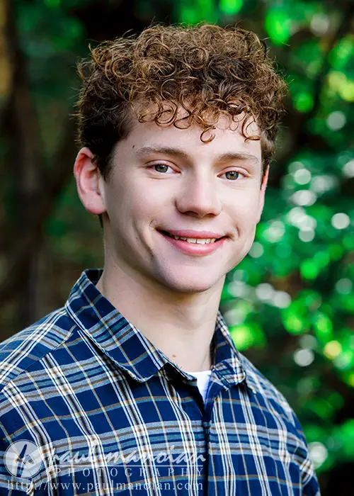 A high school senior with curly hair smiles while standing outdoors, perfect for Ann Arbor senior pictures. They are wearing a blue and white checkered shirt, and the background showcases lush greenery and blurred foliage.