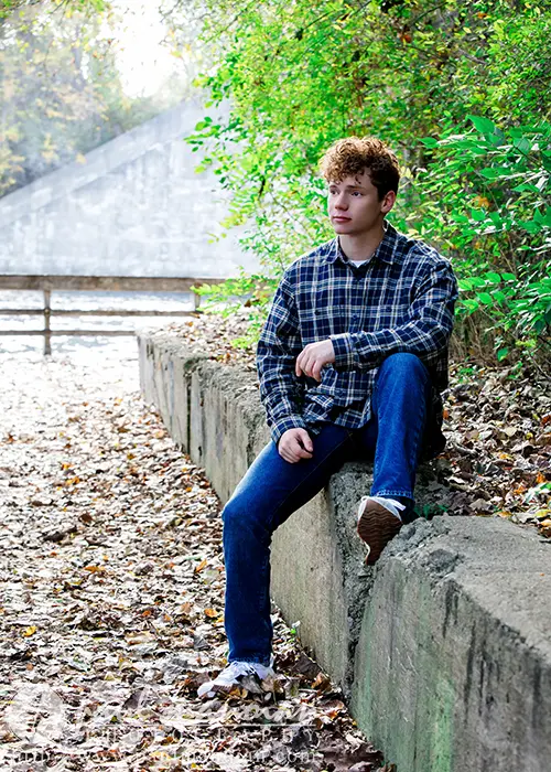 A high school senior with curly hair, perfect for Ann Arbor senior pictures, wears a plaid shirt and jeans while sitting on a concrete wall in the park. The ground is adorned with fallen leaves, and green bushes and trees provide a picturesque background.