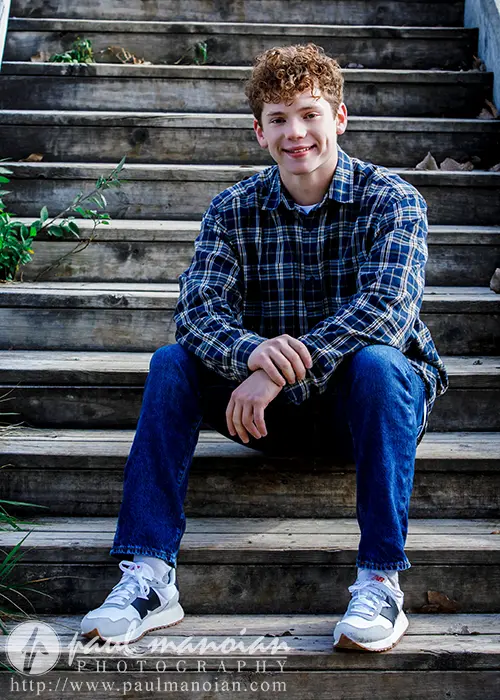 A high school senior with curly hair sits on outdoor wooden steps, capturing the essence of Ann Arbor senior pictures. Clad in a plaid shirt, jeans, and sneakers, he smiles softly with hands clasped in front. Green foliage frames the scene on the left. Photography credit is visible at the bottom.