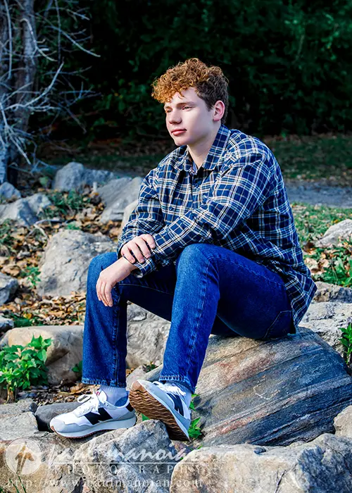 A high school senior with curly hair sits on a rock outdoors, capturing the essence of Ann Arbor senior pictures. They are wearing a plaid shirt, jeans, and sneakers, surrounded by vibrant green foliage and rustic rocks in the background.