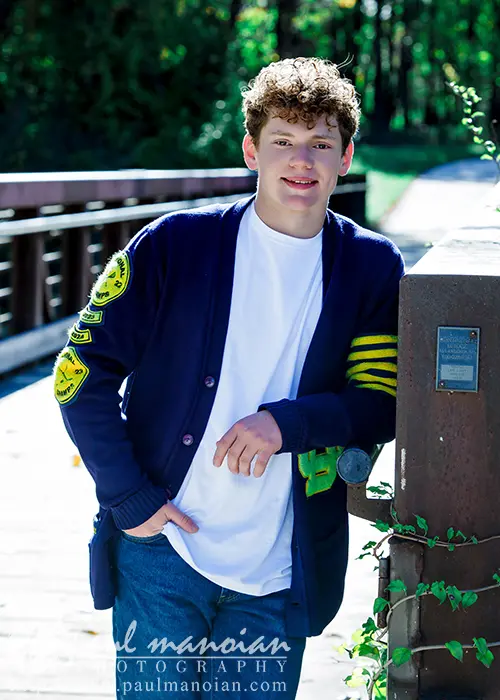 A high school senior with curly hair leans against a wooden railing on a sunny day, capturing the essence of Ann Arbor senior pictures. They wear a blue cardigan with yellow patches over a white shirt and jeans, framed by the lush green foliage in the background.