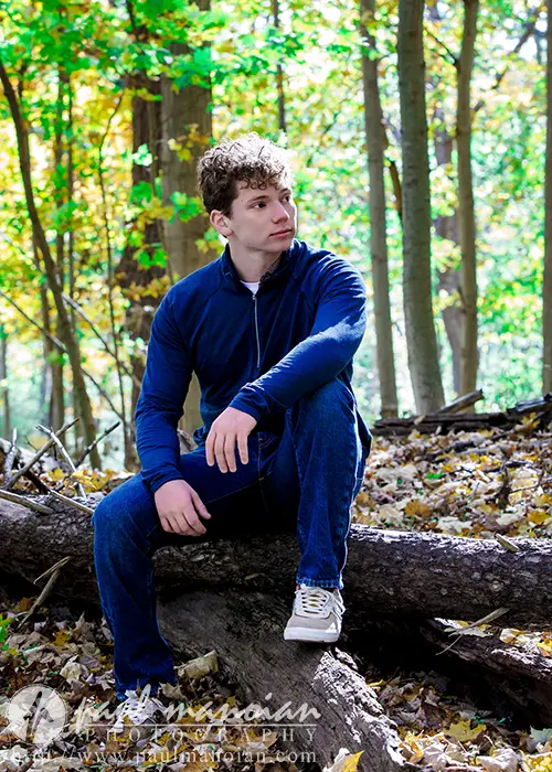 A high school senior with curly hair wearing a navy jacket and jeans is sitting on a fallen tree in the forest. Sunlight filters through the leaves, illuminating the autumn scene in Ann Arbor. The ground is carpeted with fallen leaves, perfect for capturing senior pictures.