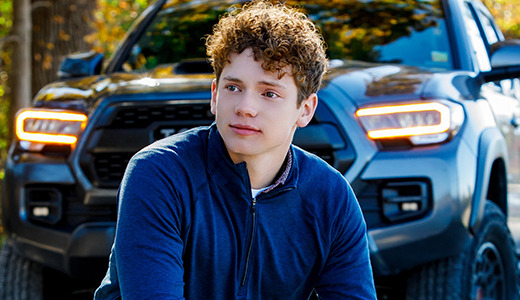 A high school senior with curly hair and a blue jacket sits on a road in front of a black pickup truck, posing for Ann Arbor senior pictures. The vibrant autumnal trees with colorful leaves create a stunning backdrop.