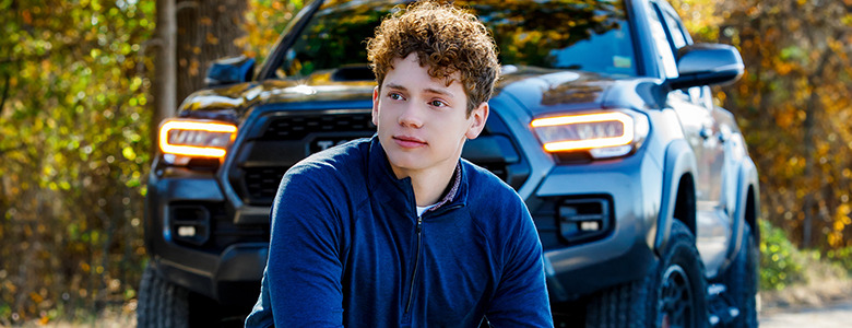 A high school senior with curly hair and a blue jacket sits on a road in front of a black pickup truck, posing for Ann Arbor senior pictures. The vibrant autumnal trees with colorful leaves create a stunning backdrop.