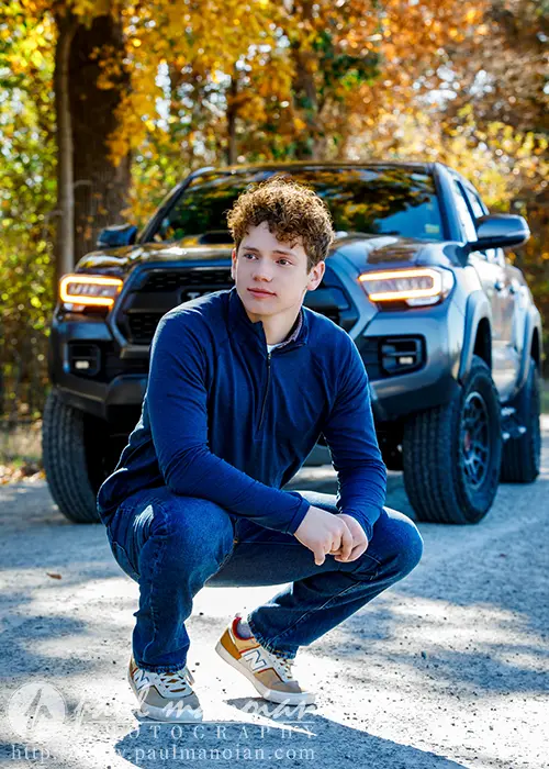 A high school senior in a blue hoodie and jeans crouches on a gravel path, striking a pose for their Ann Arbor senior pictures in front of a black pickup truck. The scene is beautifully framed by trees adorned with vibrant autumn foliage.
