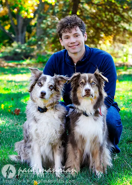 In Ann Arbor, a high school senior kneels on grass for their senior pictures, smiling at the camera as two brown and white dogs sit in front. The background showcases trees adorned with vibrant autumn foliage.