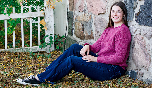 A high school senior in a pink sweater and jeans sits on the ground leaning against a stone wall. She is smiling and surrounded by fallen leaves, with a white fence and green foliage in the background.