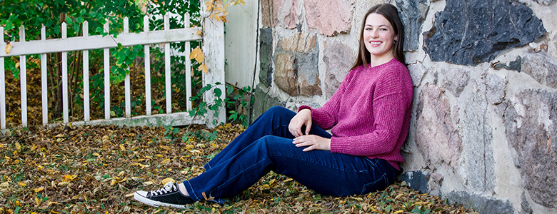 A high school senior in a pink sweater and jeans sits on the ground leaning against a stone wall. She is smiling and surrounded by fallen leaves, with a white fence and green foliage in the background.