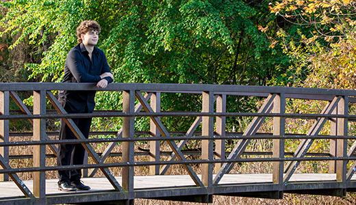 A high school senior with curly hair stands on a wooden bridge, gazing into the distance. The background is filled with lush green trees and some foliage showing autumn colors. The scene is serene and peaceful.