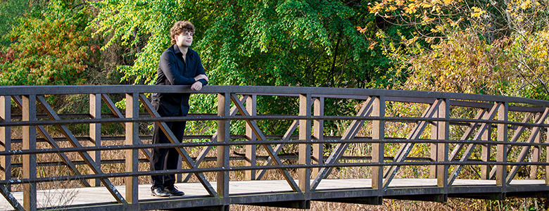 A high school senior with curly hair stands on a wooden bridge, gazing into the distance. The background is filled with lush green trees and some foliage showing autumn colors. The scene is serene and peaceful.