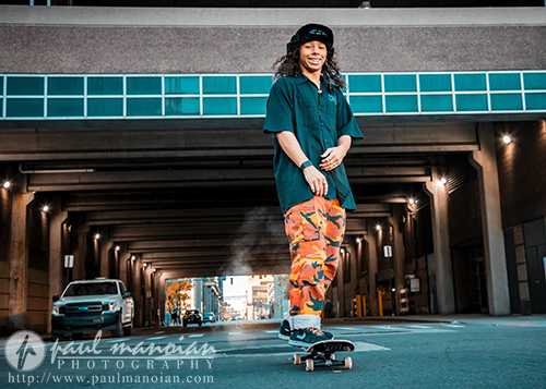 A high school senior skateboards confidently in an urban tunnel, capturing the essence of energetic senior pictures. They sport a dark shirt, orange camouflage pants, and a black hat. A car lingers in the backdrop, while the lighting enhances this dynamic street scene.