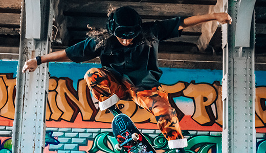 A skateboarder in mid-air performs a trick under a bridge, their skateboard flipping beneath them. They wear a black hat, a dark shirt, and orange patterned pants. The background bursts with colorful graffiti art on concrete pillars, adding an edgy flair perfect for senior pictures.