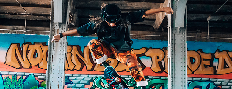 A skateboarder in mid-air performs a trick under a bridge, their skateboard flipping beneath them. They wear a black hat, a dark shirt, and orange patterned pants. The background bursts with colorful graffiti art on concrete pillars, adding an edgy flair perfect for senior pictures.