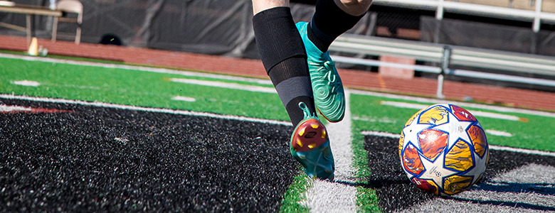 A soccer player in black socks and teal cleats is poised to kick a colorful ball on a green field, while capturing dynamic senior pictures. The scene features benches and a running track in the background.