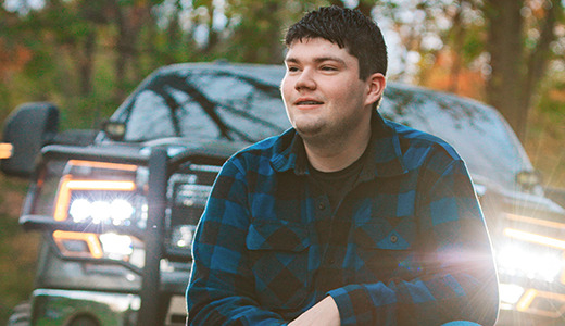 A high school senior wearing a blue and black plaid shirt is sitting in front of a truck with bright headlights on, surrounded by a forested area. The scene conveys a sense of tranquility in nature.