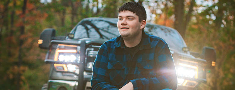 A high school senior wearing a blue and black plaid shirt is sitting in front of a truck with bright headlights on, surrounded by a forested area. The scene conveys a sense of tranquility in nature.