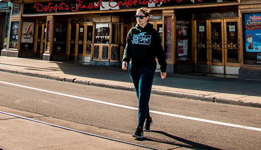 A high school senior wearing sunglasses and a black jacket strolls down an empty street in Allen Park, where sunlight dances on the pavement in front of a theater. It's the perfect backdrop for capturing senior pictures.