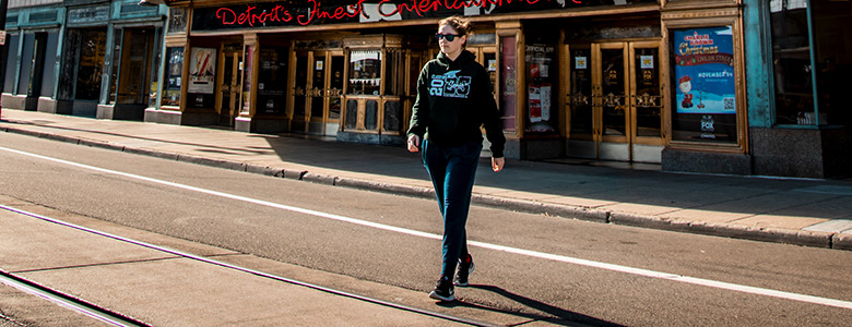 A high school senior wearing sunglasses and a black jacket strolls down an empty street in Allen Park, where sunlight dances on the pavement in front of a theater. It's the perfect backdrop for capturing senior pictures.