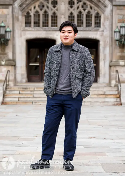 A high school senior stands confidently on a stone path in front of a historic-looking building with ornate windows, capturing the perfect Ann Arbor senior picture moment. Dressed in a gray jacket and dark pants, they look directly towards the camera.