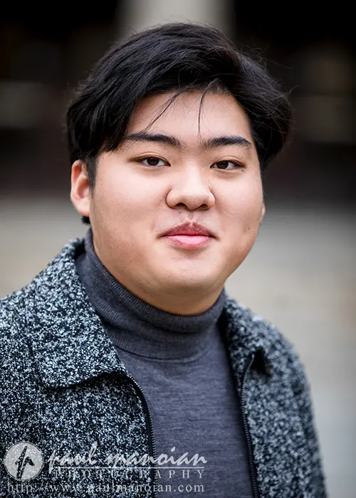 A high school senior with short dark hair, wearing a gray turtleneck and a textured coat, gazes at the camera with a neutral expression. The softly blurred background gives an elegant touch, reminiscent of Ann Arbor senior pictures.