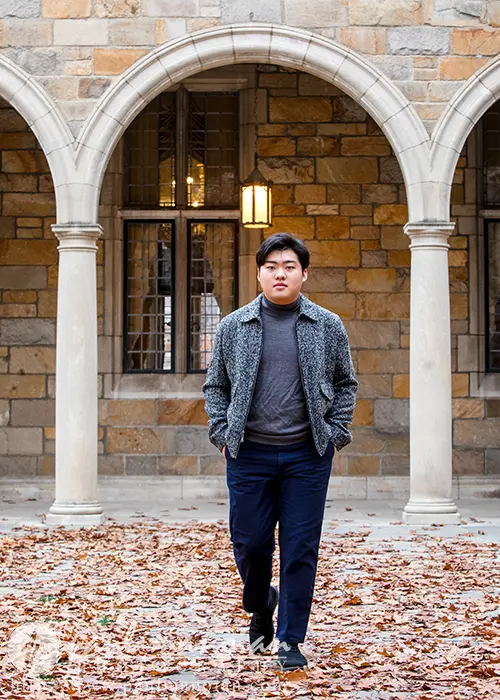 A high school senior confidently walks in front of a stone building with arched columns, capturing the essence of Ann Arbor senior pictures. The ground is covered with fallen autumn leaves, and the individual sports a patterned jacket and dark pants.