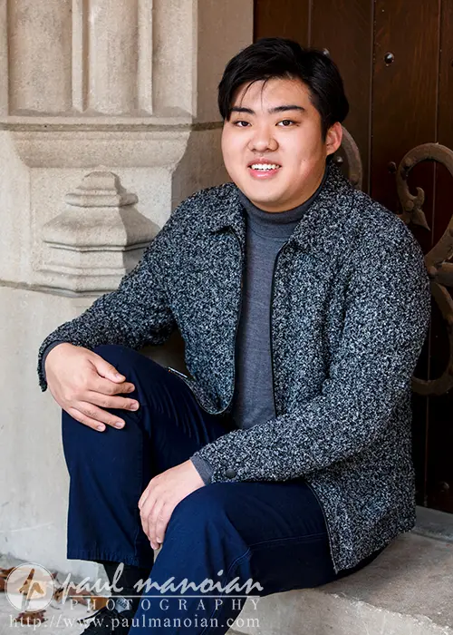 A high school senior with short dark hair sits on stone steps in front of a wooden door, wearing a grey sweater and dark pants. They are smiling with their hands resting on their knees, capturing the essence of Ann Arbor senior pictures through this candid moment.