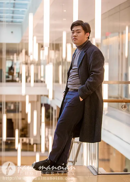 A high school senior wearing a dark coat and pants, with a gray turtleneck, leans casually against a glass railing in a modern indoor setting in Ann Arbor. Bright lighting fixtures hang from the ceiling, creating an ideal backdrop for senior pictures.