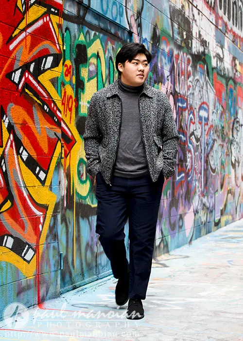 A high school senior walks confidently down a vibrant graffiti-covered alley in Ann Arbor, capturing memories for their senior pictures. They wear a gray jacket and dark pants, blending seamlessly into the artistic, colorful background.