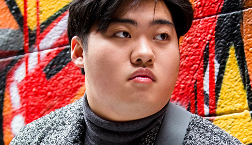 A high school senior with dark hair gazes sideways, posing for their Ann Arbor senior pictures against a vibrant graffiti wall splashed with red, orange, black, and yellow hues. They sport a gray patterned jacket over a dark turtleneck.