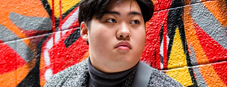 A high school senior with dark hair gazes sideways, posing for their Ann Arbor senior pictures against a vibrant graffiti wall splashed with red, orange, black, and yellow hues. They sport a gray patterned jacket over a dark turtleneck.