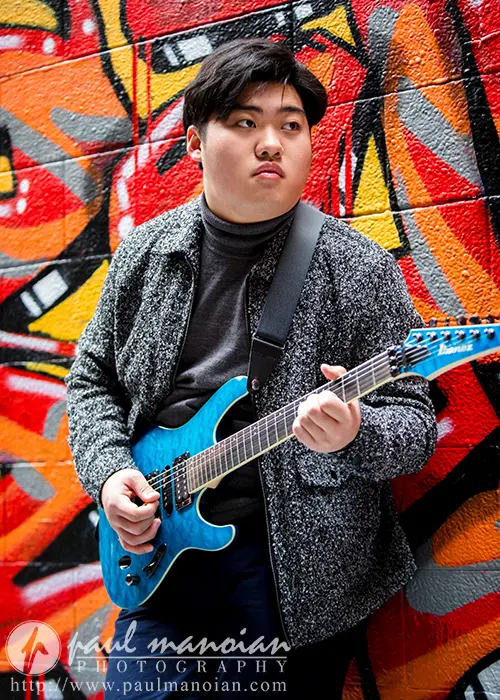 In front of a colorful graffiti wall, a high school senior playing a blue electric guitar captures the unique vibe typical of Ann Arbor senior pictures. Dressed in a gray jacket and black shirt, they glance thoughtfully to the side, embodying creativity and style.
