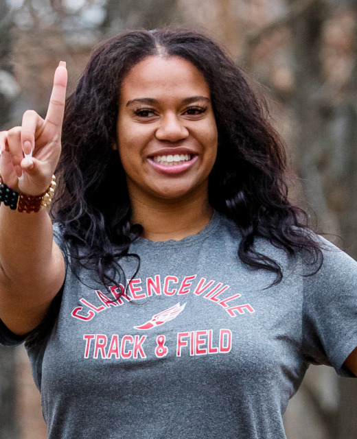 A high school senior smiling and holding up one finger in a wooded area. They are wearing a "Glencoeville Track & Field" t-shirt.