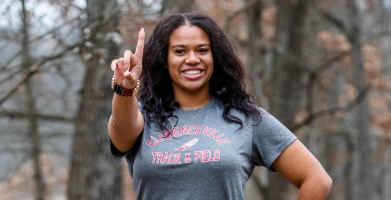 A high school senior smiling and holding up one finger in a wooded area. They are wearing a "Glencoeville Track & Field" t-shirt.