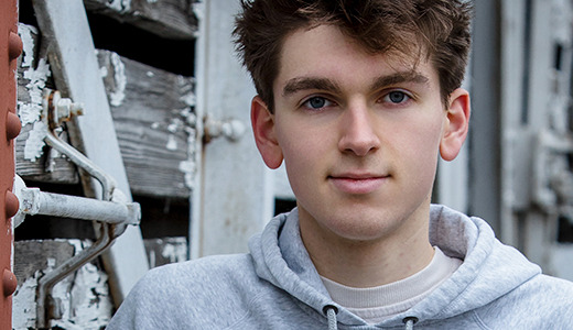 A high school senior with short, tousled hair, wearing a gray hoodie and posing for his Divine Child senior pictures, stands in front of a weathered wooden structure with peeling paint. He looks directly at the camera as metal elements and a rust-colored section complete the background.