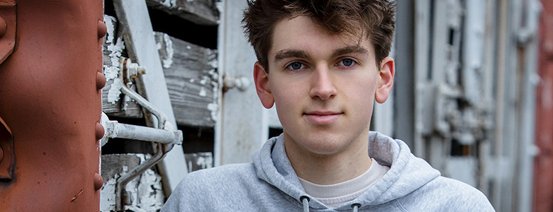 A high school senior with short, tousled hair, wearing a gray hoodie and posing for his Divine Child senior pictures, stands in front of a weathered wooden structure with peeling paint. He looks directly at the camera as metal elements and a rust-colored section complete the background.