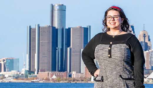 A high school senior stands smiling in front of a body of water, with a city skyline in the background for their Divine Child senior portraits session. The skyline includes several tall buildings and a clear blue sky.
