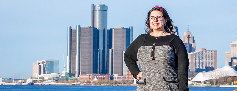 A high school senior stands smiling in front of a body of water, with a city skyline in the background for their Divine Child senior portraits session. The skyline includes several tall buildings and a clear blue sky.