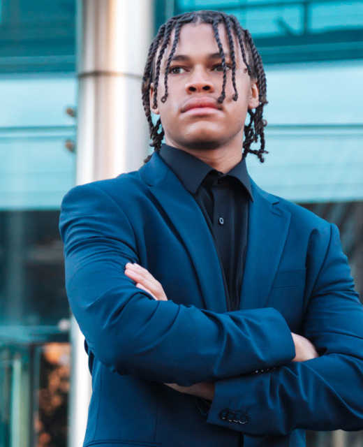 A high school senior with braided hair stands confidently with arms crossed, wearing a dark suit. They are in front of a modern glass building, exuding a professional and determined demeanor.