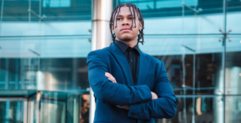 A high school senior with braided hair stands confidently with arms crossed, wearing a dark suit. They are in front of a modern glass building, exuding a professional and determined demeanor.