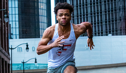 A high school senior in athletic gear sprints down a downtown Detroit street, perfect for vibrant senior pictures. He wears a white tank top with a logo and gray shorts. Tall buildings and a street sign labeled "Larned" are visible in the background.